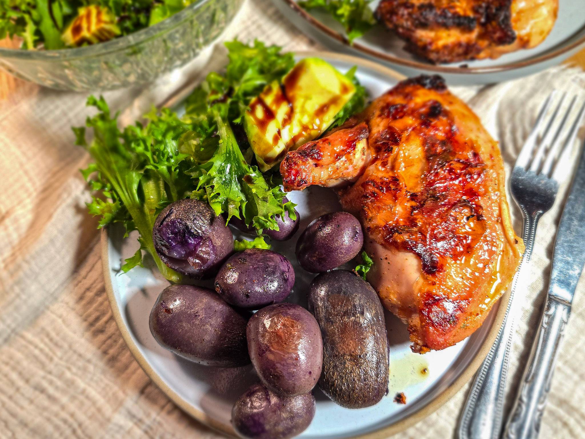A plate of purple potatoes with grilled chicken supreme and a green salad with avocado and balsamic glaze.