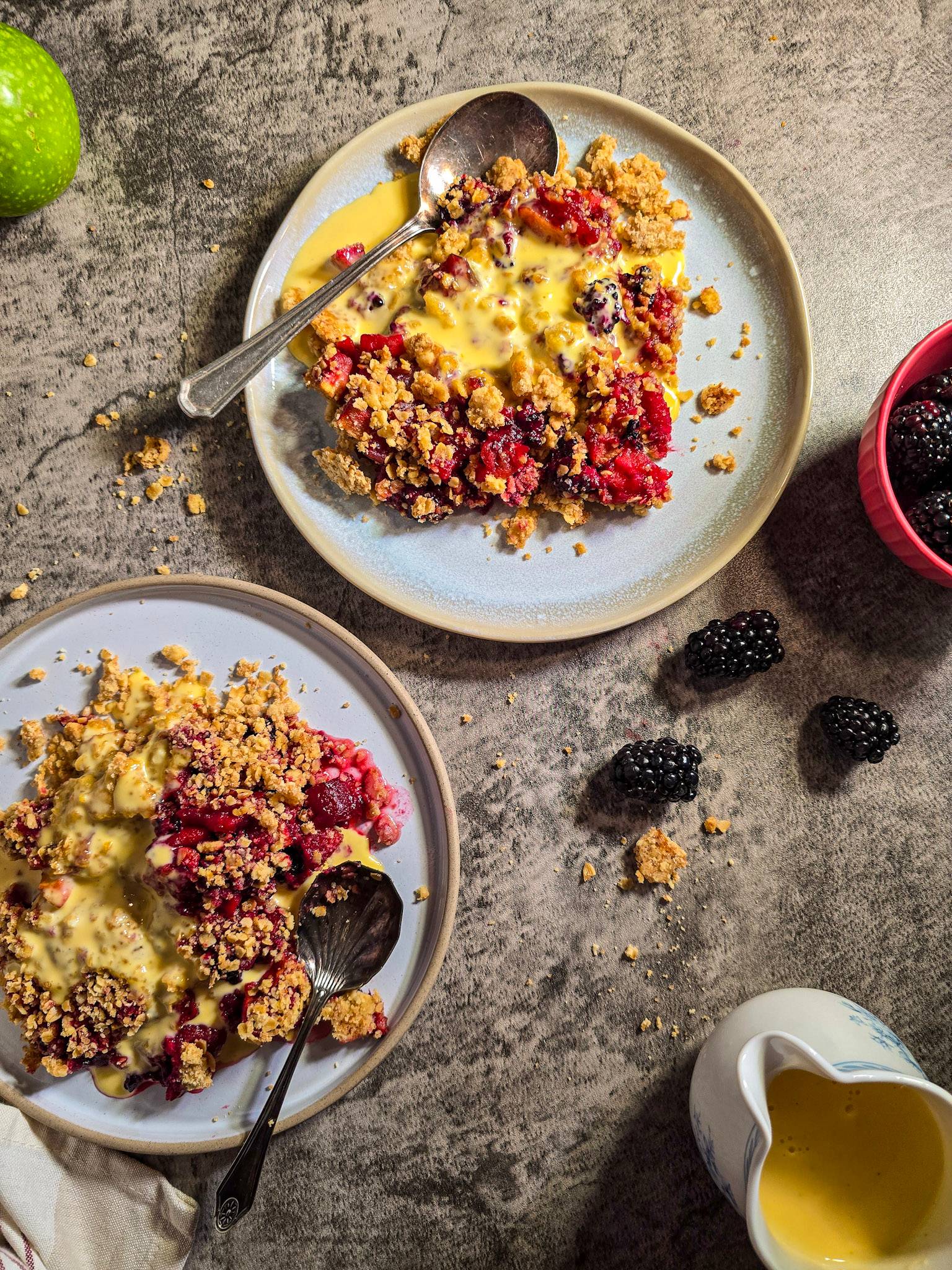 A close-up top-down view of apple and blackberry crumble served on two plates with custard, extra blackberries scattered around and a small jug of custard.