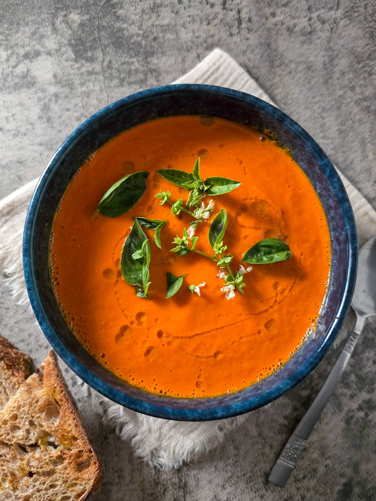Smooth tomato soup topped with basil leaves and olive oil in a blue bowl.