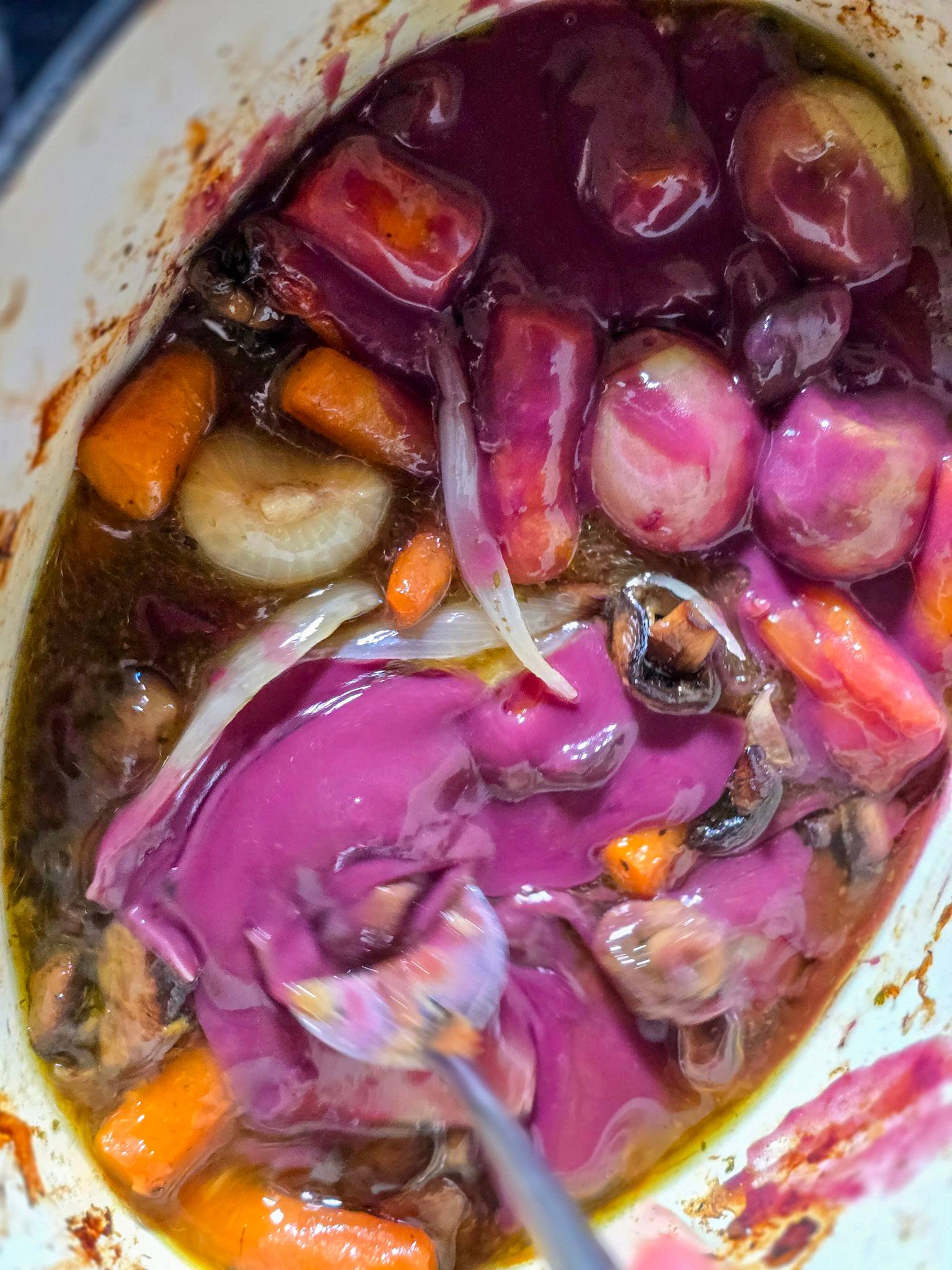Close-up of red wine gravy being stirred around roasted carrots, onions and mushrooms in a casserole dish, glossy purple sauce swirling through the vegetables.