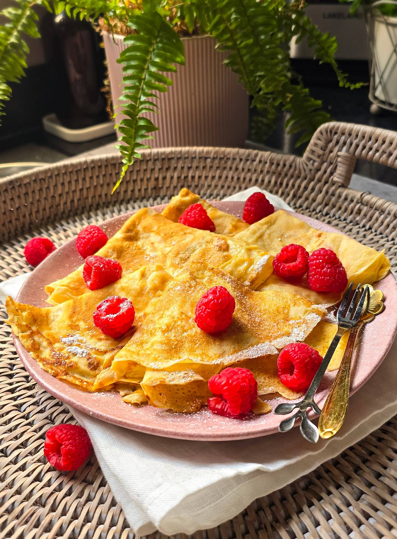 Golden buttermilk crepes folded on a pink plate, dusted with icing sugar and topped with fresh raspberries.