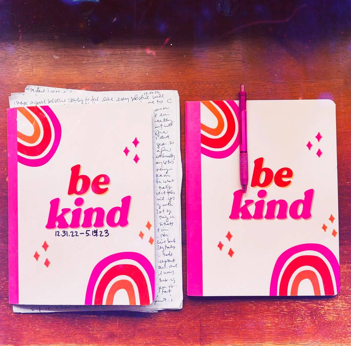 two pink notebooks on a brown desk. Text reads be kind. 