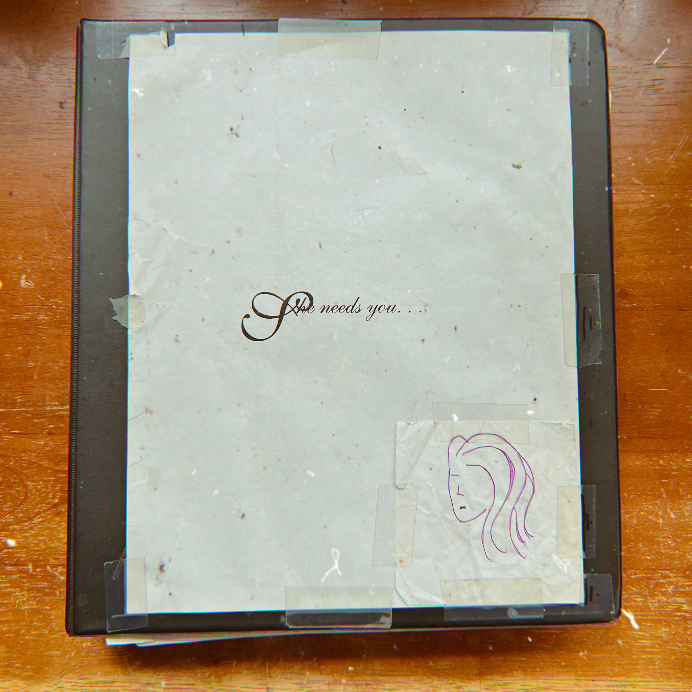 black spiral binder on brown desk. Text on binder reads, "She needs you." Bottom right hand corner is a drawing of a female face with hair. 