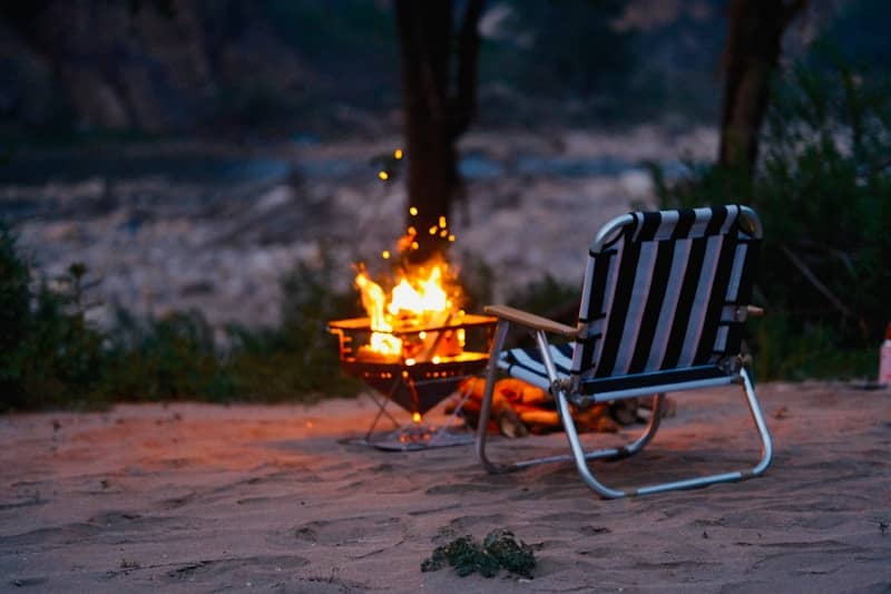 A striped chair sits by a campfire at dusk.