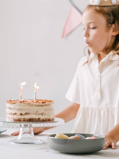 A young girl in a white dress blowing out birthday candles at a celebration indoors.