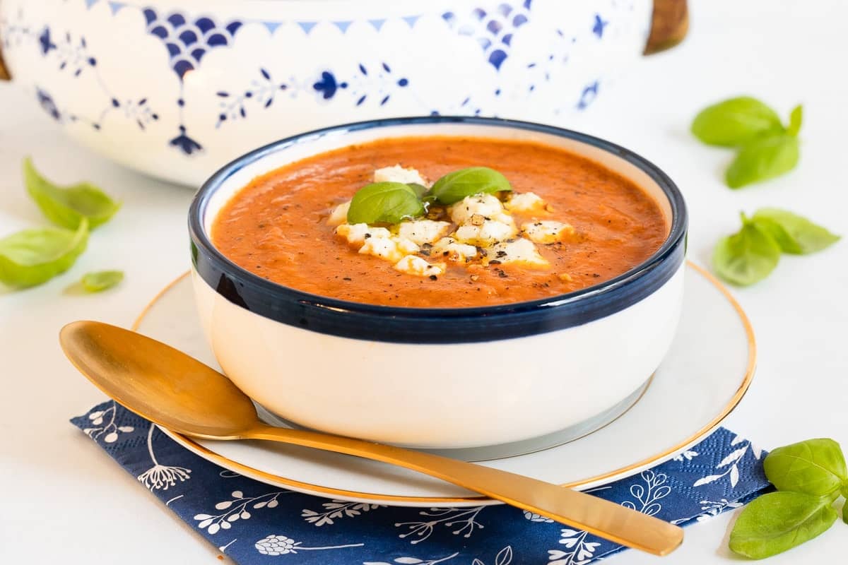 White bean and tomato basil soup with cheese and fresh basil in a blue and white bowl with a gold spoon.