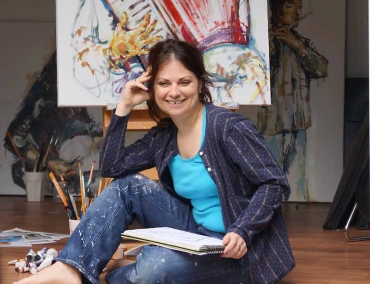 Woman artist Paola Minekov sits on the floor holding a sketchbook, smiling, with art supplies around her and a colorful painting on an easel in the background.