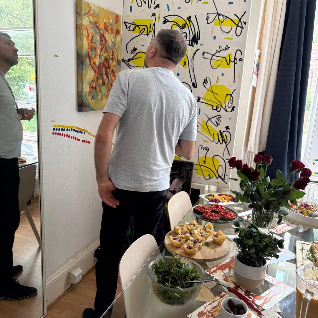 A man stands in front of abstract artwork by artist Paola Minekov, with a table set with various dishes, roses, and a salad in the foreground.