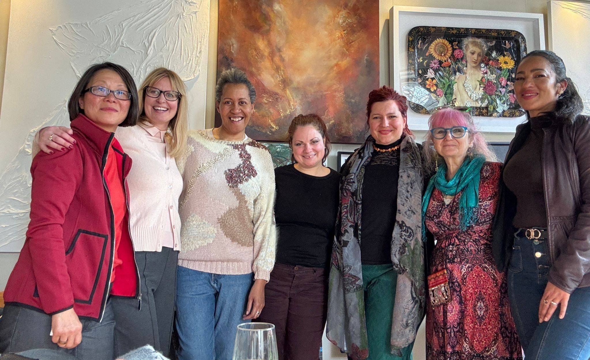 Seven women stand side by side indoors, smiling at the camera, with art pieces displayed on the wall behind them.