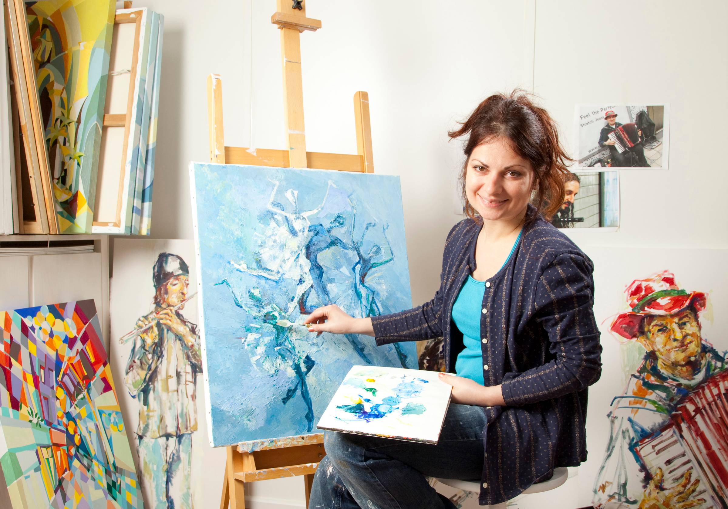 Female artist Paola Minekov sits on a stool painting a blue-toned abstract tree on a canvas, surrounded by colorful paintings and art supplies in a studio.