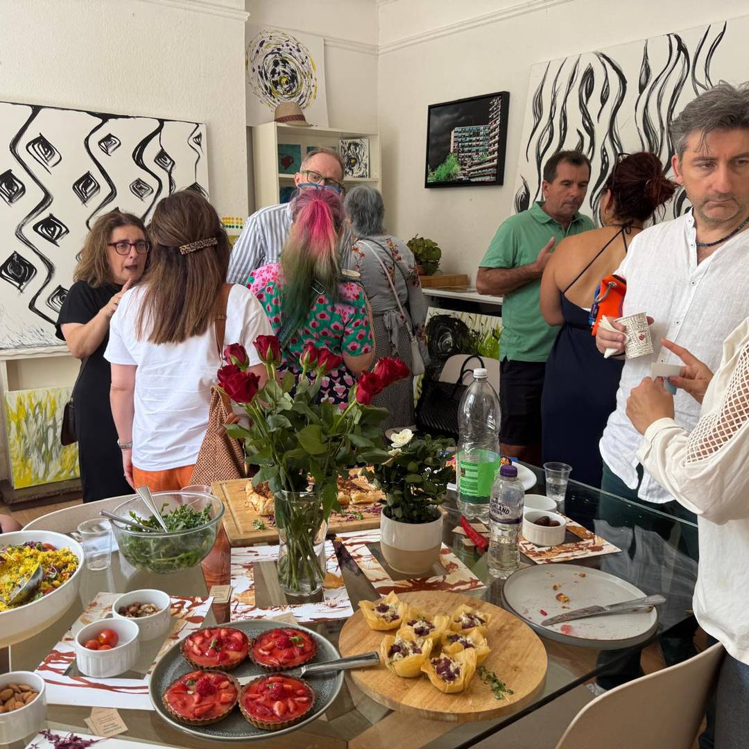 A group of people socialize around a table with various foods and drinks in a room decorated with abstract artwork and flowers.
