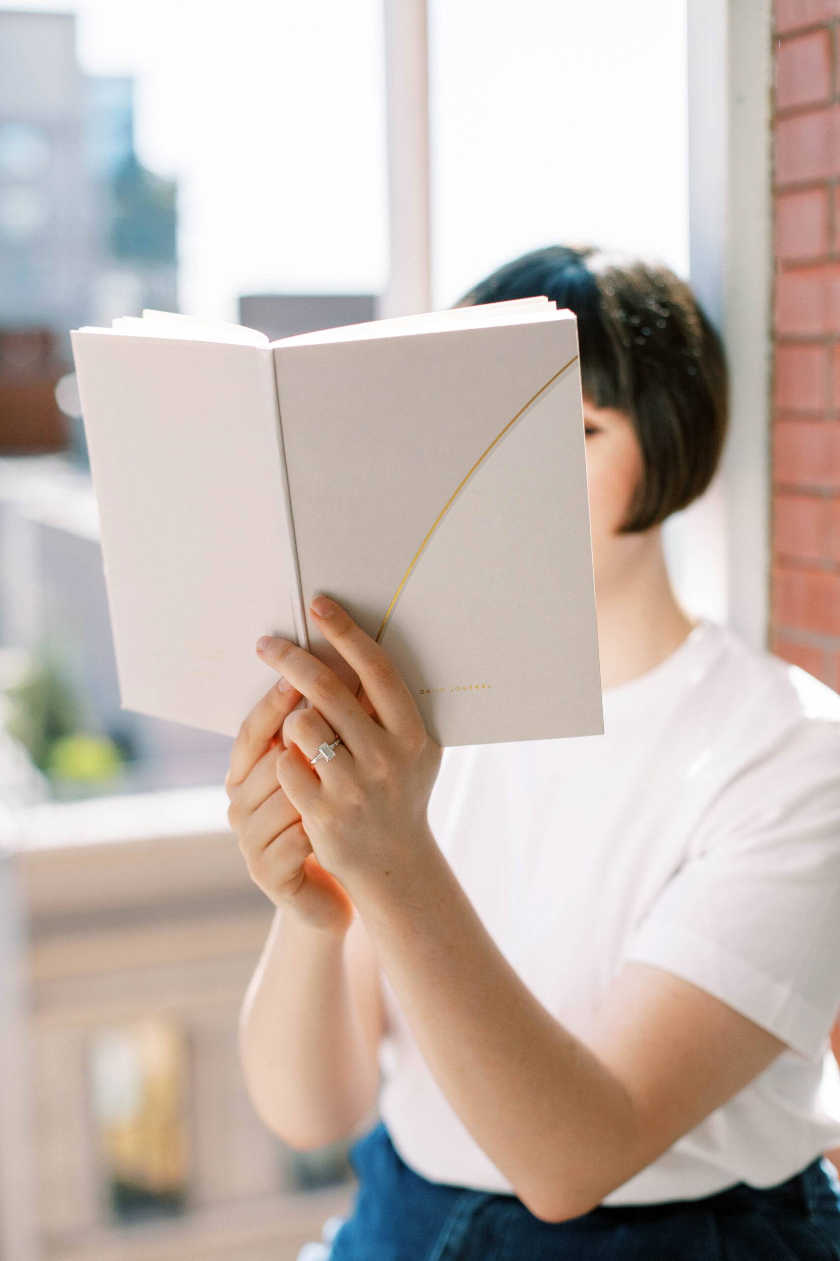 Woman seated by the window is reading a book.