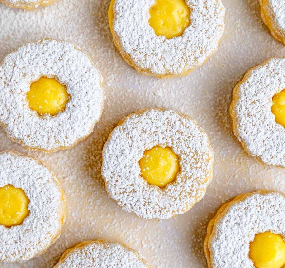 Delicate sandwich cookies dusted with powdered sugar are arranged neatly. Each cookie has a central opening filled with smooth yellow lemon curd, creating a visually appealing contrast with the white sugar coating. The cookies are placed on a light backgr