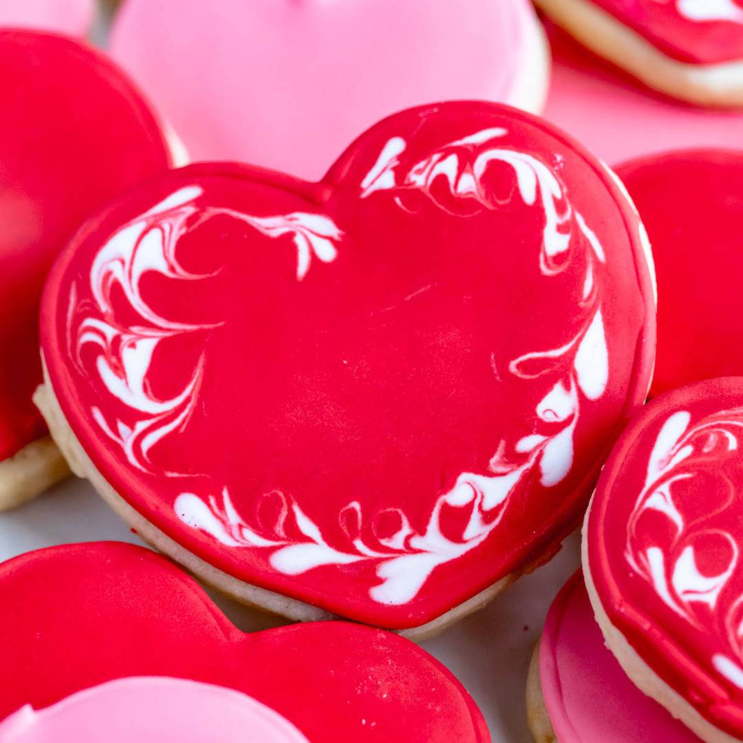 A close-up of heart-shaped cookies decorated with bright red and pink icing. The central cookie features intricate white swirls on the red icing, creating a decorative pattern. Other cookies in the background are solid shades of red and pink.