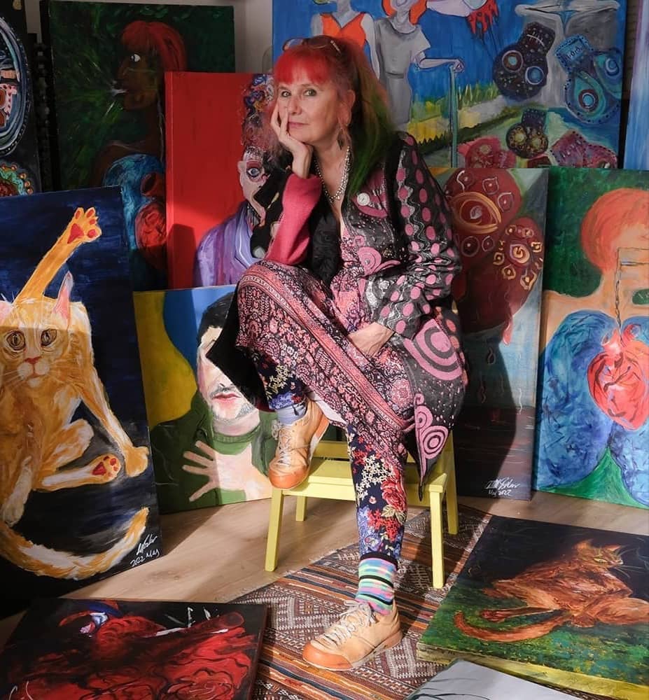 Contemporary female artist Michelle Baharier, with red hair, wearing patterned clothing, sits surrounded by her vibrant paintings and surreal fine art in an art studio.