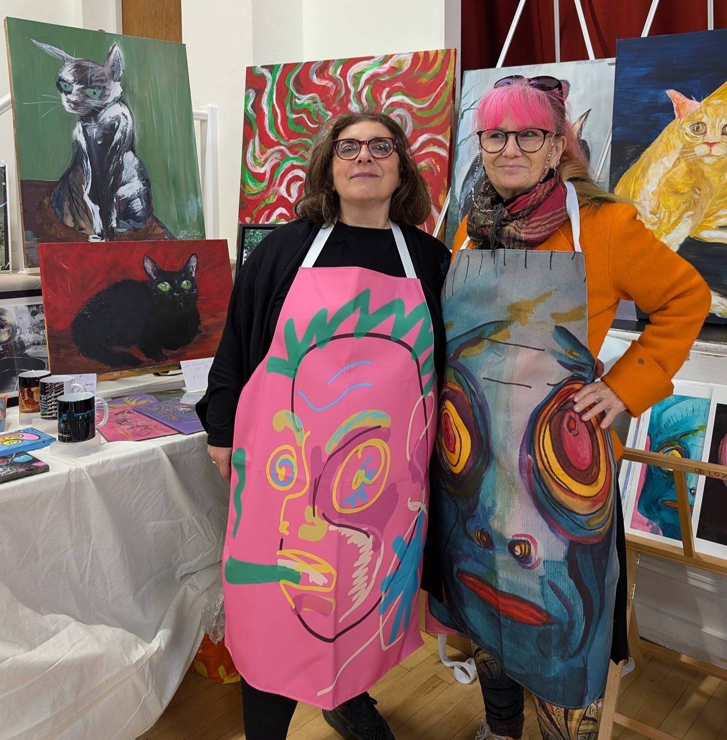 Two women artists wearing colourful aprons stand in front of a display of cat-themed and abstract paintings at an indoor art exhibition