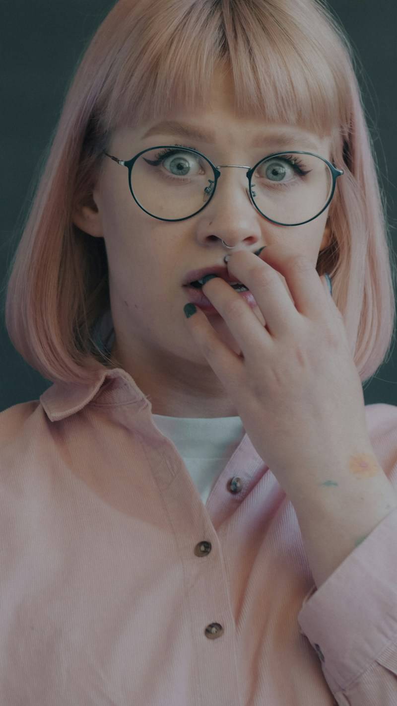 A person with pink hair and glasses biting their fingernails in a pink shirt
