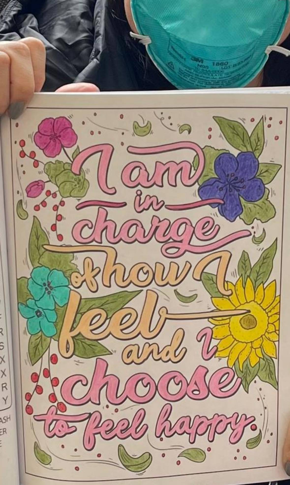 A person wearing a teal face mask holds a colorful page with the quote "I am in charge of how I feel and I choose to feel happy." The page features decorative flowers, including pink, blue, and purple blooms along with a yellow sunflower, surrounded by gr