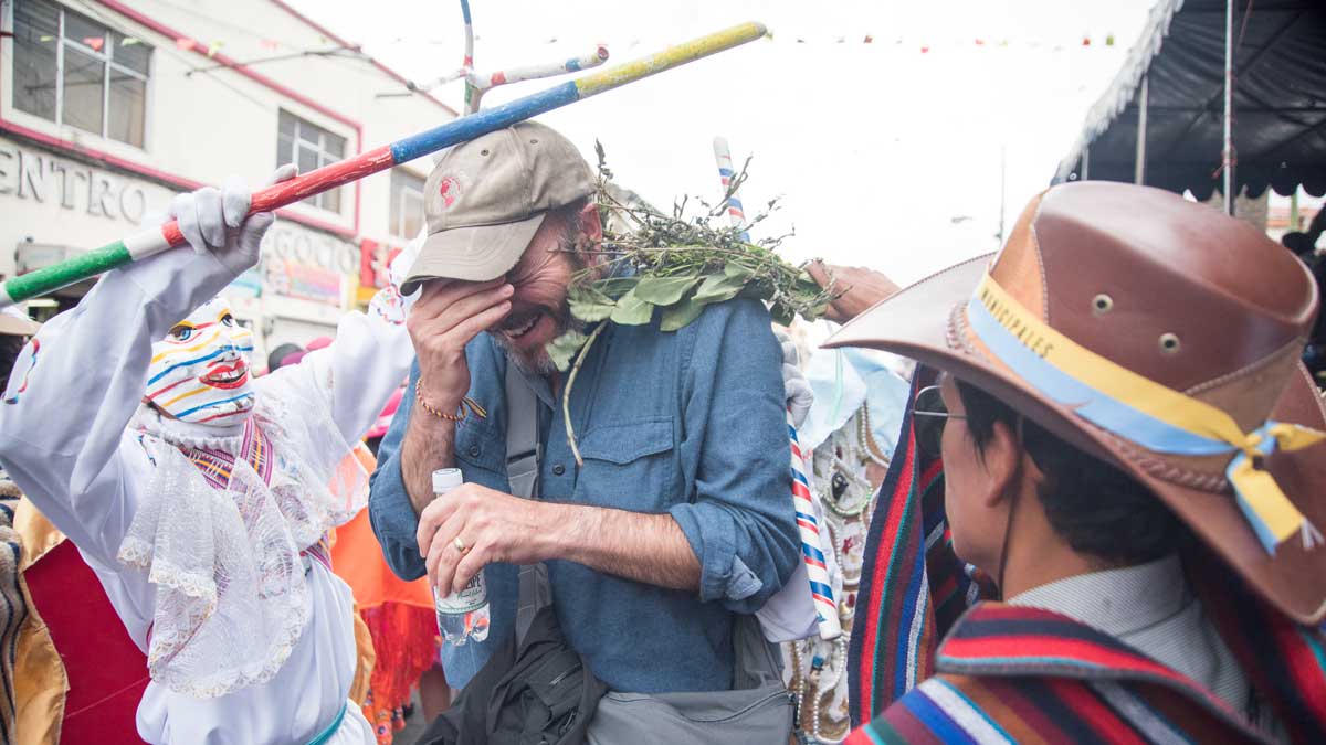 A tourist covers his face with one hand while costumed figures touch him with sticks and branches, blessing him during the Mama Negra parade in Latacunga