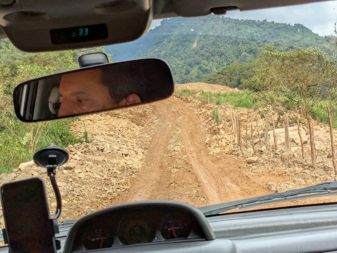 Looking through the front windshield of the car, the view is a rutted, muddy dirt road leading towards forested mountains.