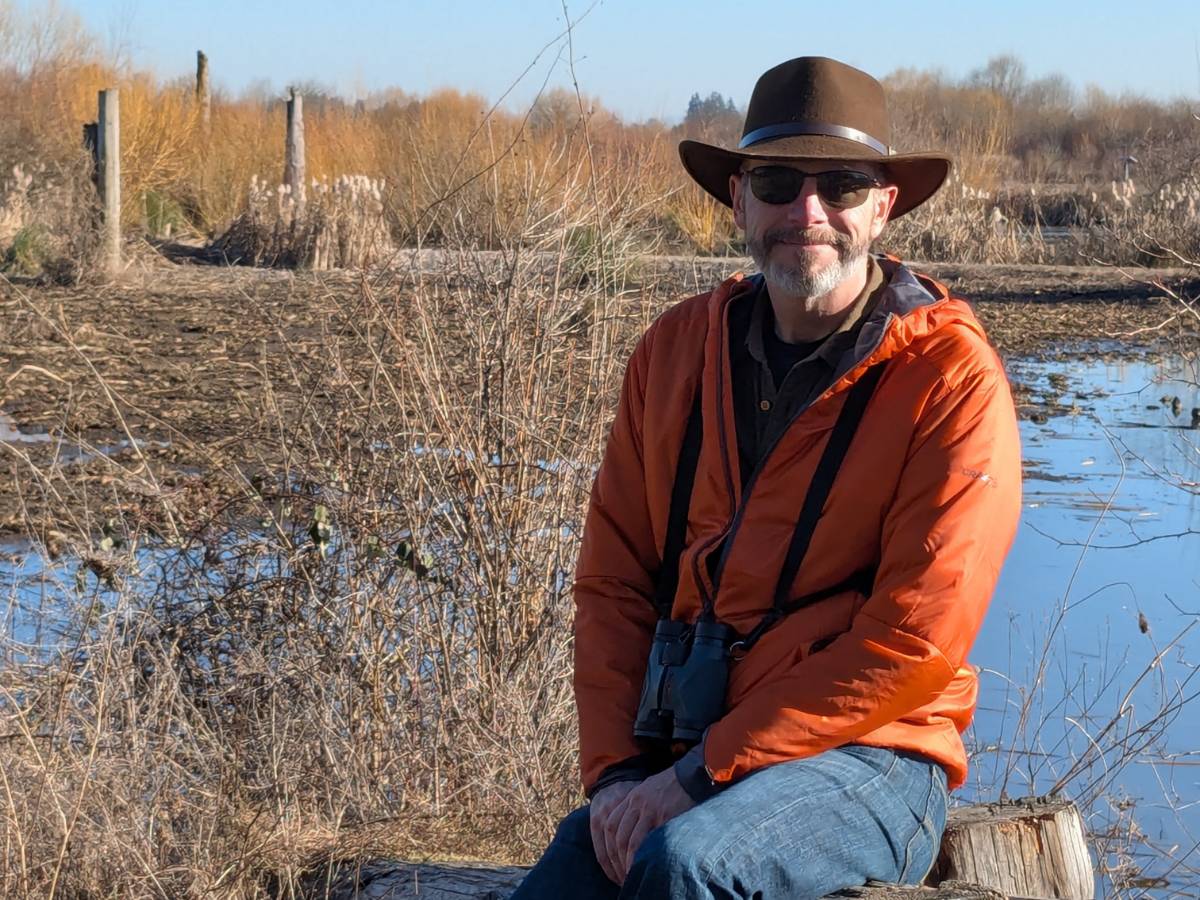 A man in an orange jacket and brown, woolen hat sits on a log near a wetlands.
