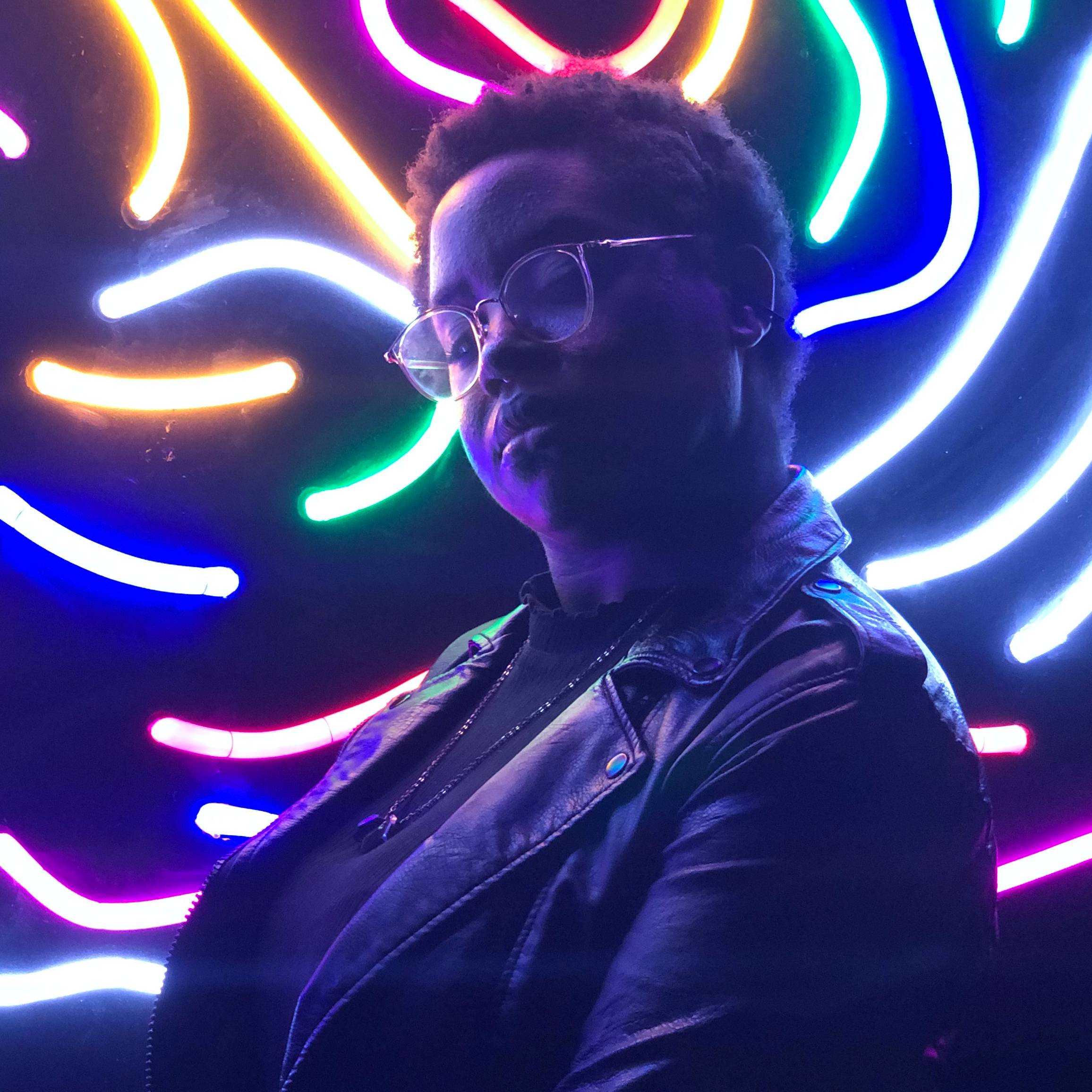 A photo of me, looking softly at the camera against a neon light art display.