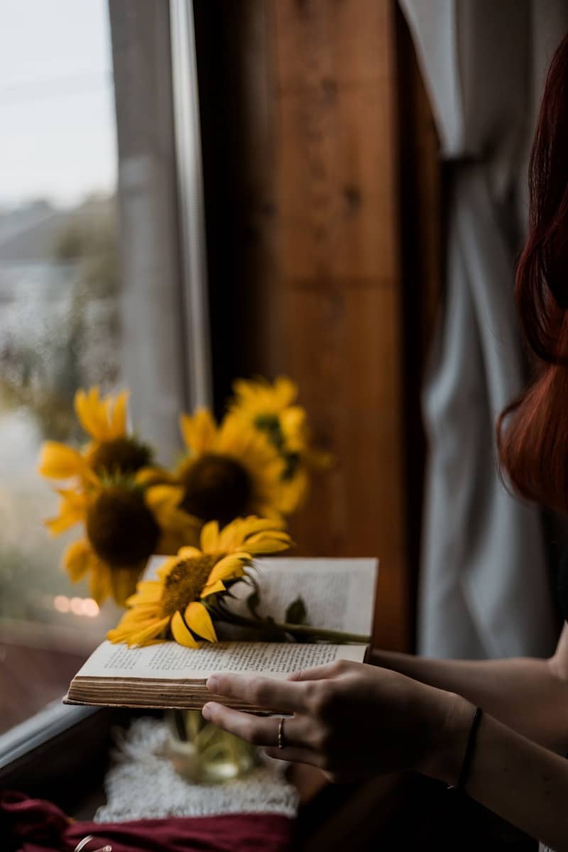 A woman reading a book in front of a window with sunflowers