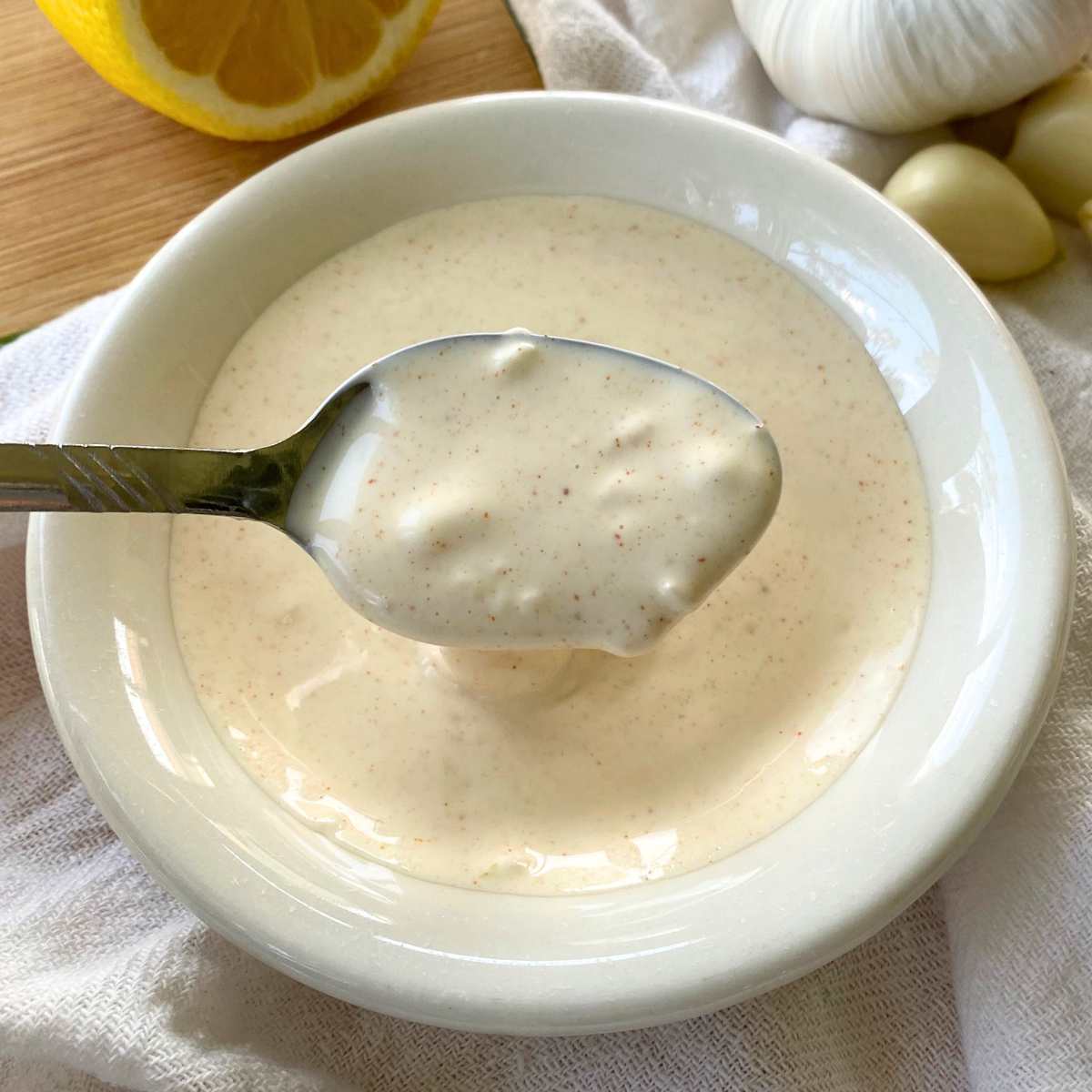 Spoon holding lemon garlic aioli over a white bowl with more.