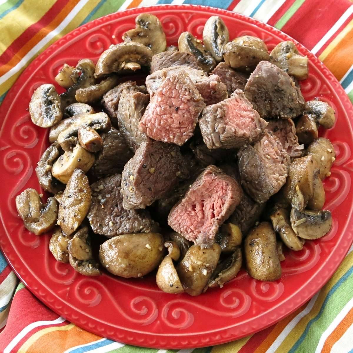 Steak bites and mushrooms on a red plate.