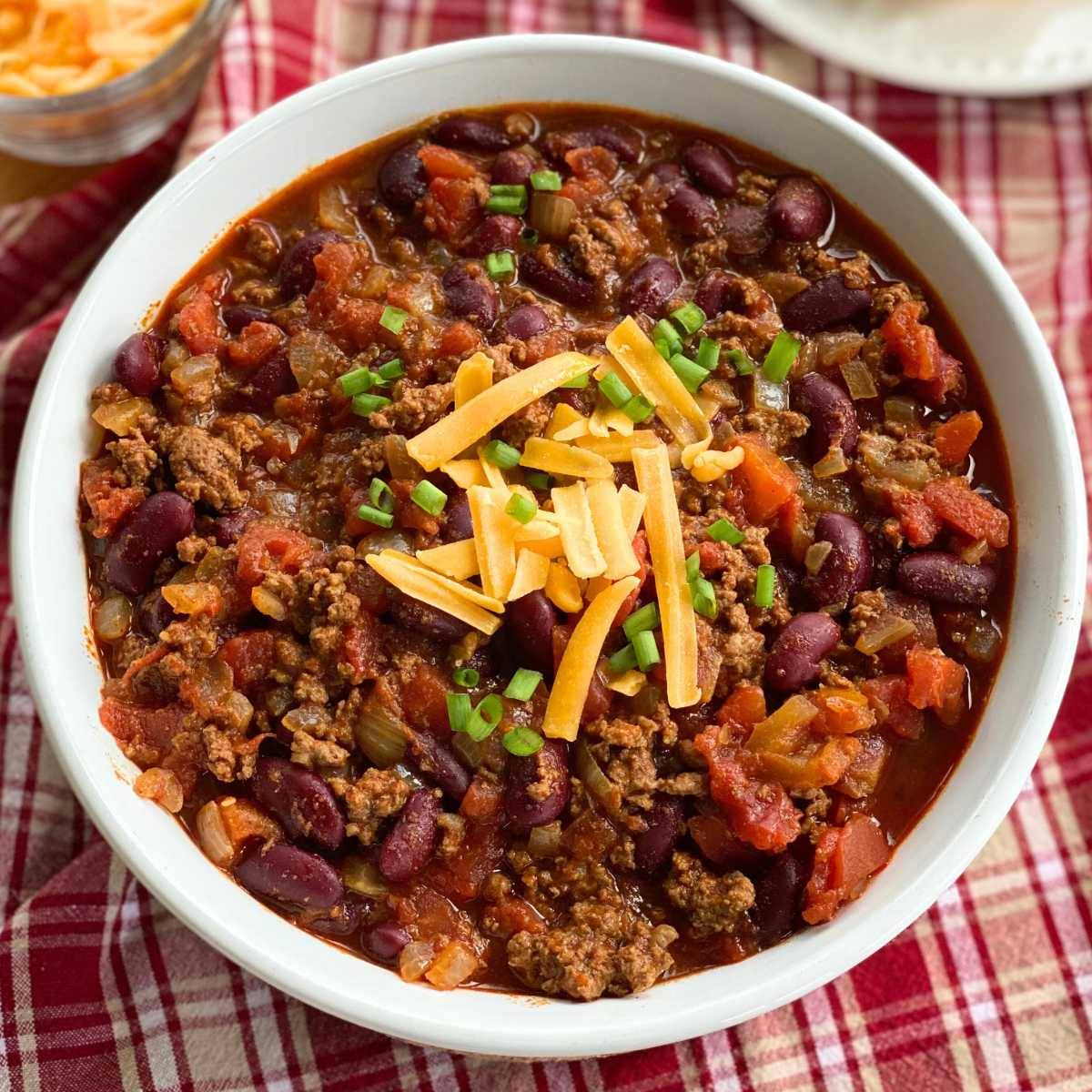 White bowl with beef and bean chili and topped with cheddar cheese and green onions.