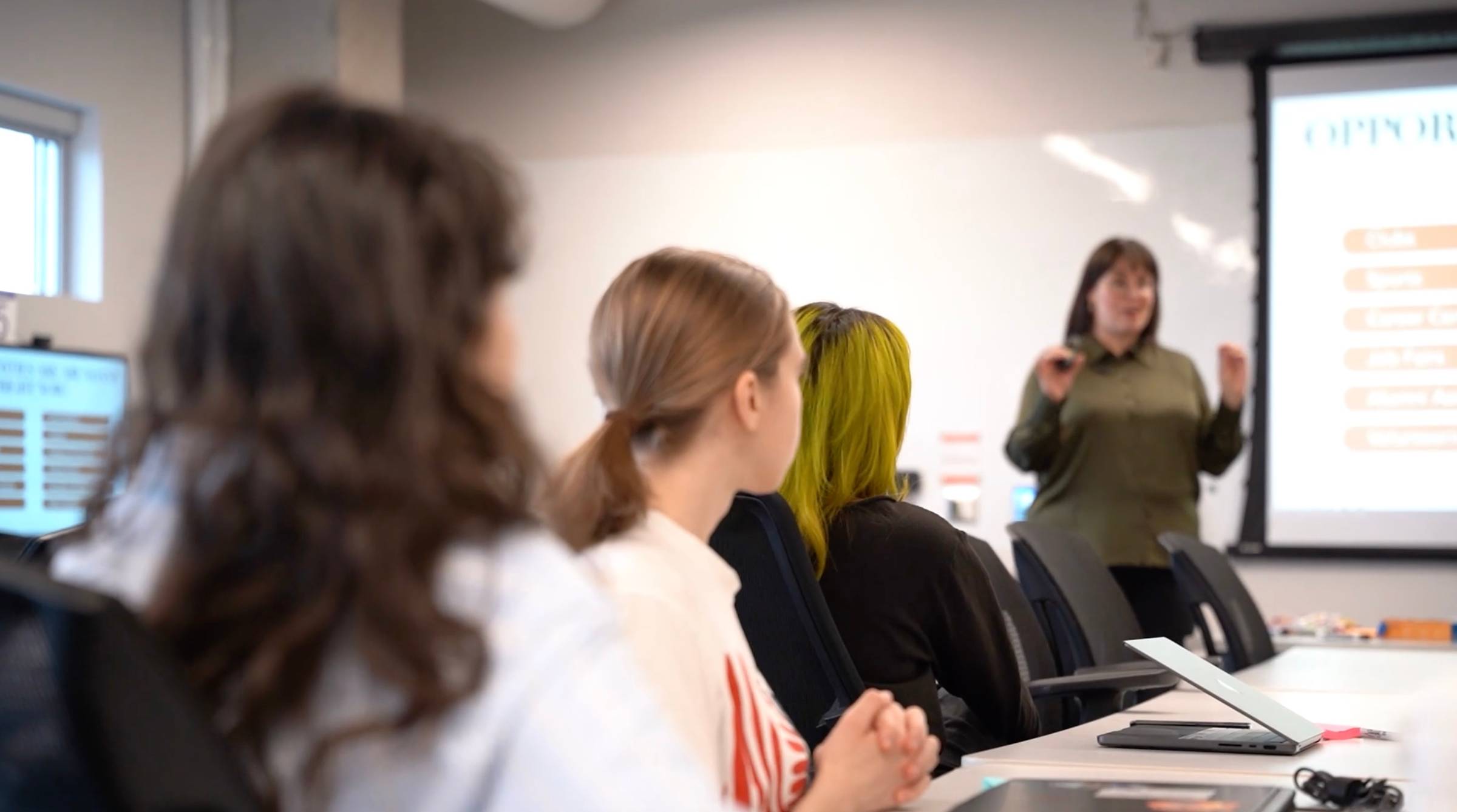 Trina Boos, facilitator gestures while speaking to a seated group of students in a classroom, seen from behind the row of attentive listeners.