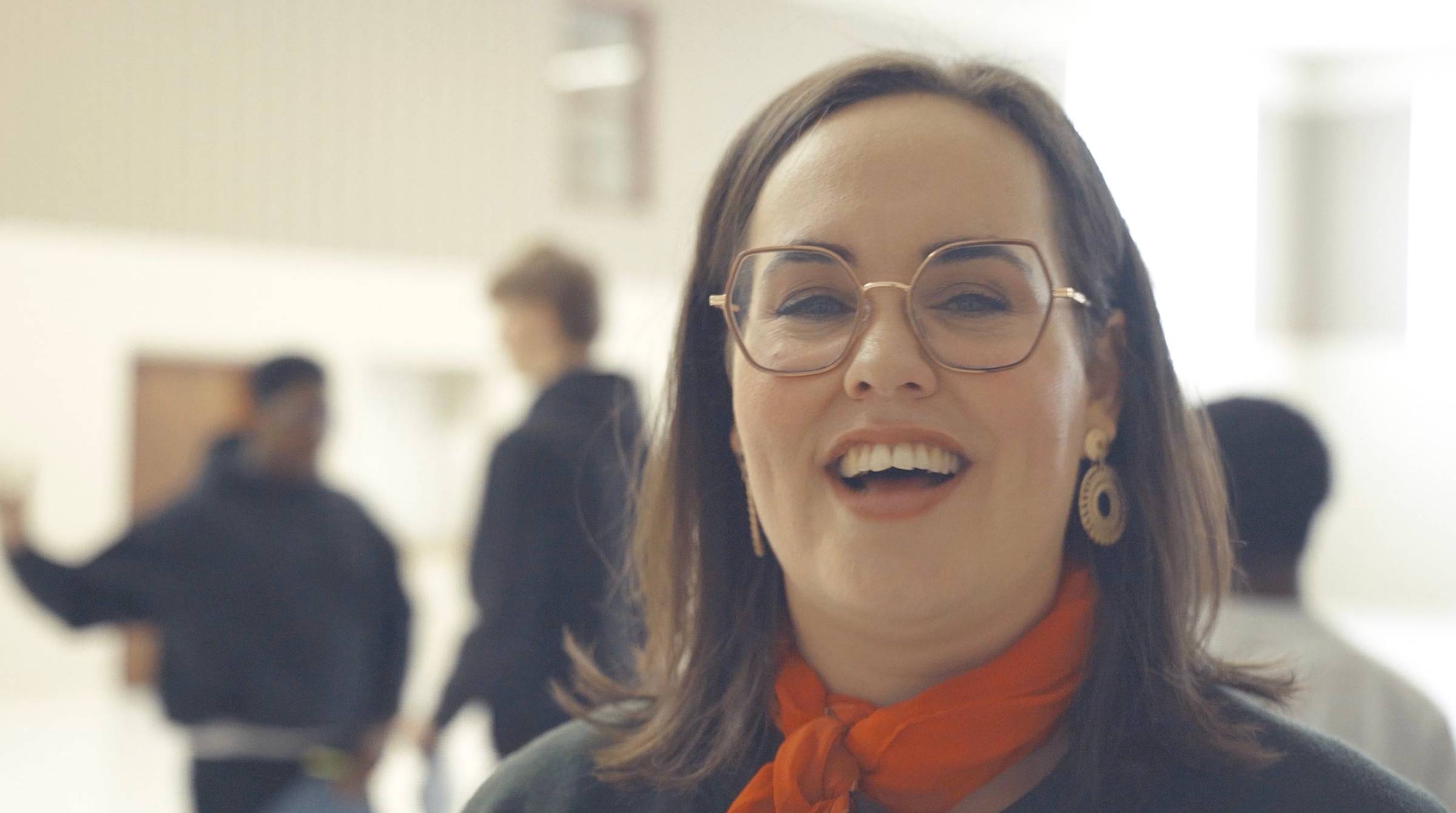 A woman with glasses and an orange scarf smiling and laughing, with a blurred group of people in the background.