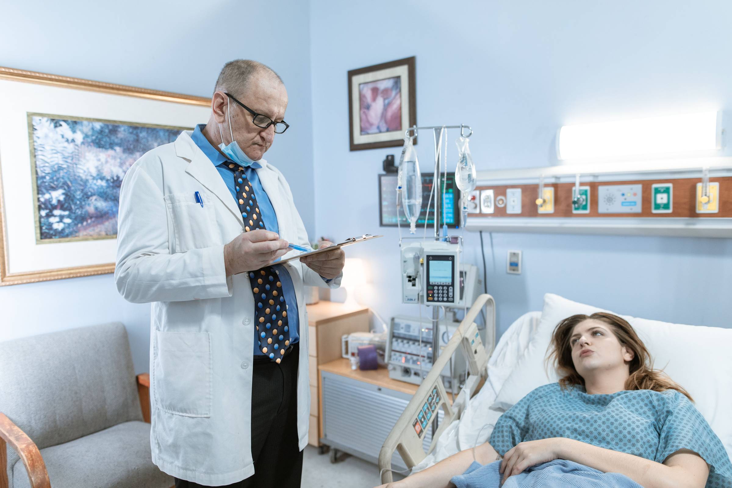 A doctor in a white coat reviews notes on a clipboard while speaking with a patient resting in a hospital bed, IV equipment and medical monitors visible beside her.
