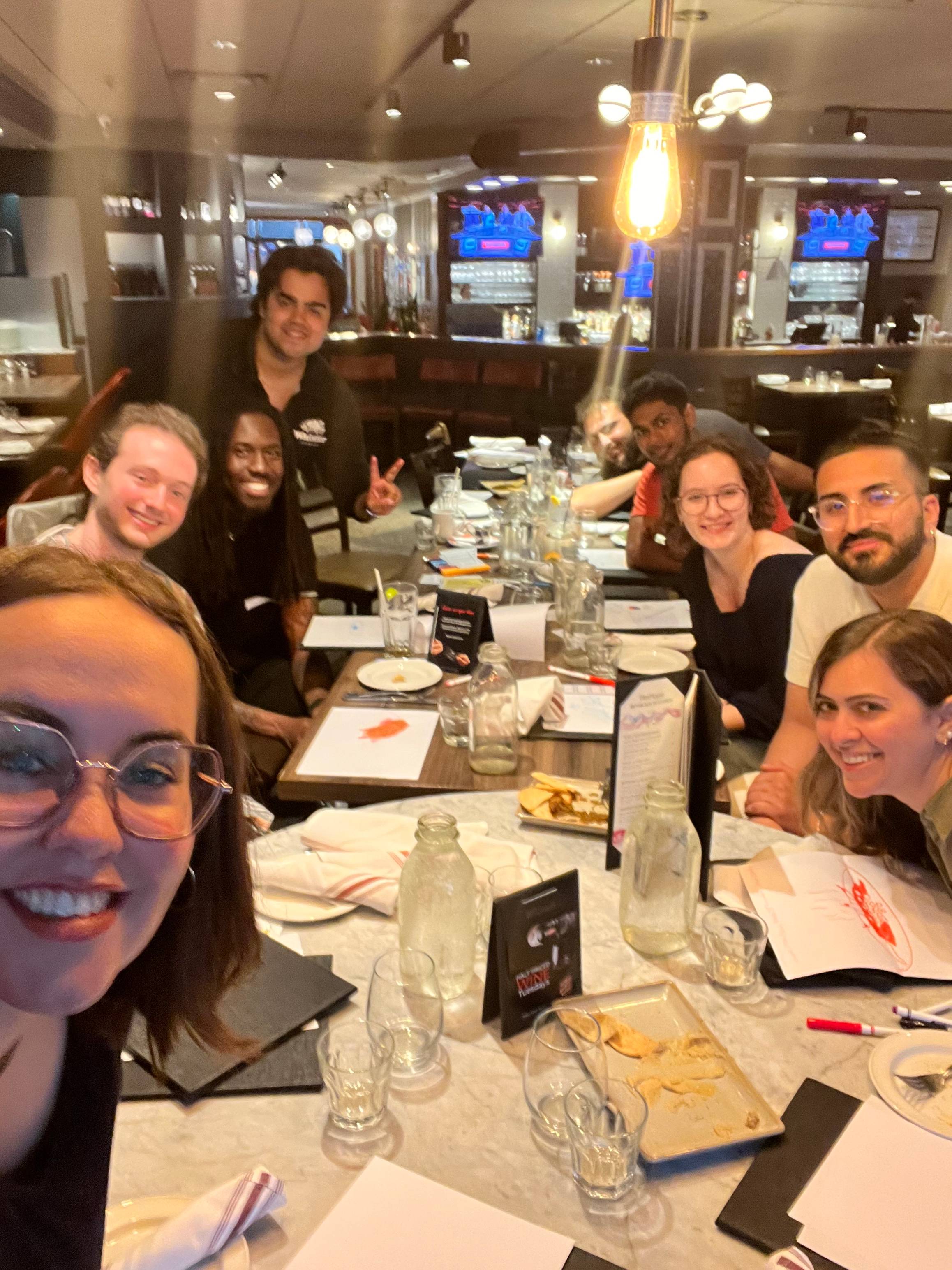 A group of about nine people gathered around a long restaurant table, smiling and posing for a selfie-style group photo. The table is set with glasses, plates, and menus, and the setting appears to be a dimly lit restaurant with wooden decor.