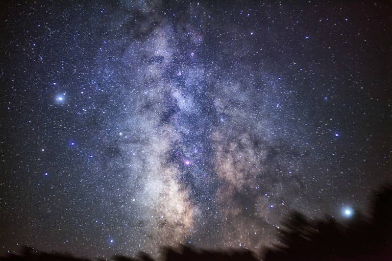 The Milky Way galaxy glowing with dense star clusters above a dark tree line