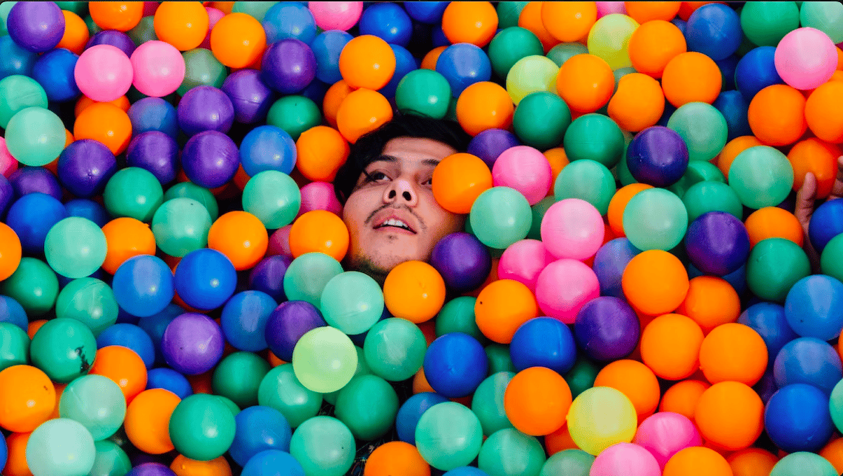 A face peers out from a ton of small multicolored plastic balls (inside a ball pit). 
