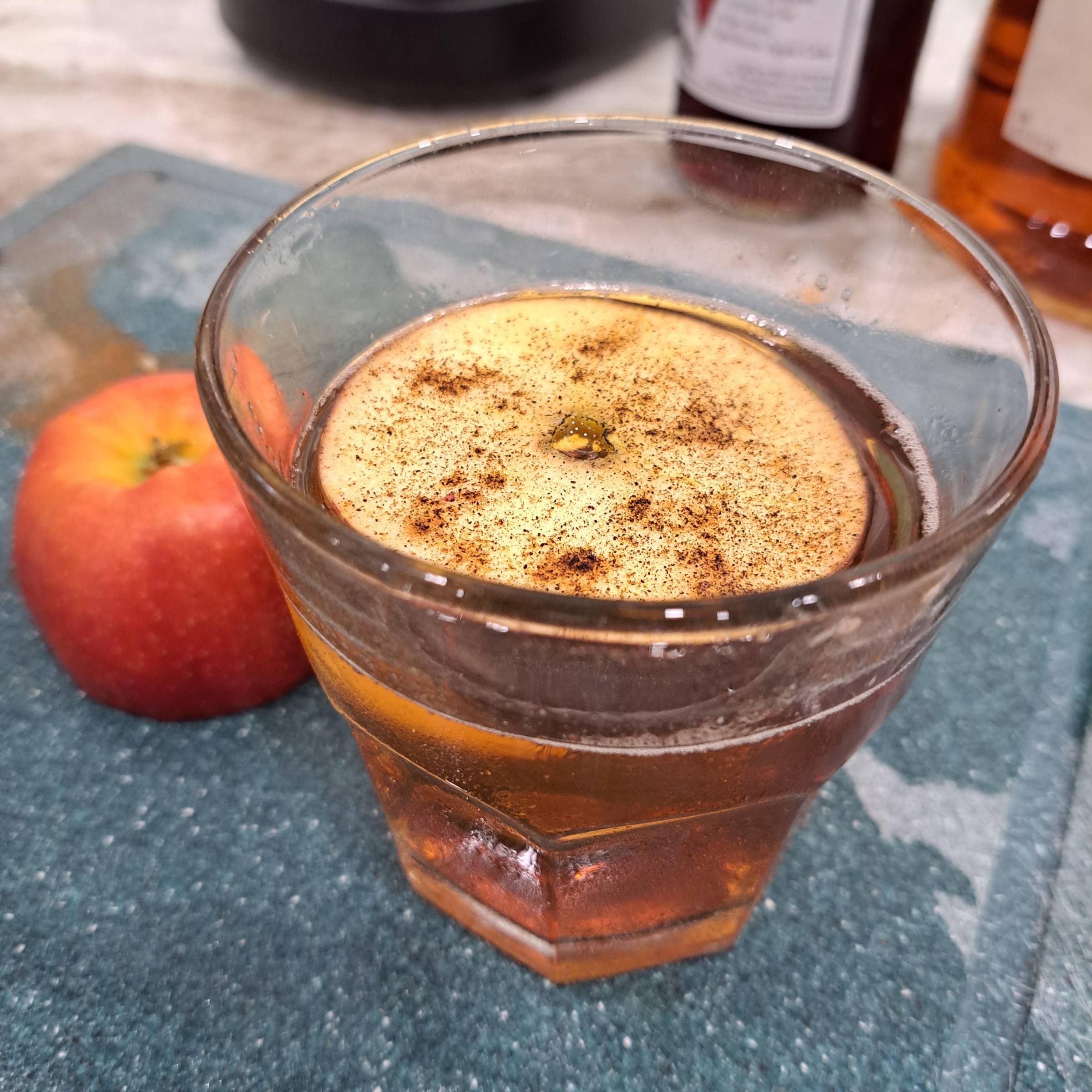 A bourbon and Calvados cocktail served over a large ice cube in a faceted rocks glass, topped with a torched apple wheel dusted with spice. A whole apple sits beside the glass on a textured blue surface.