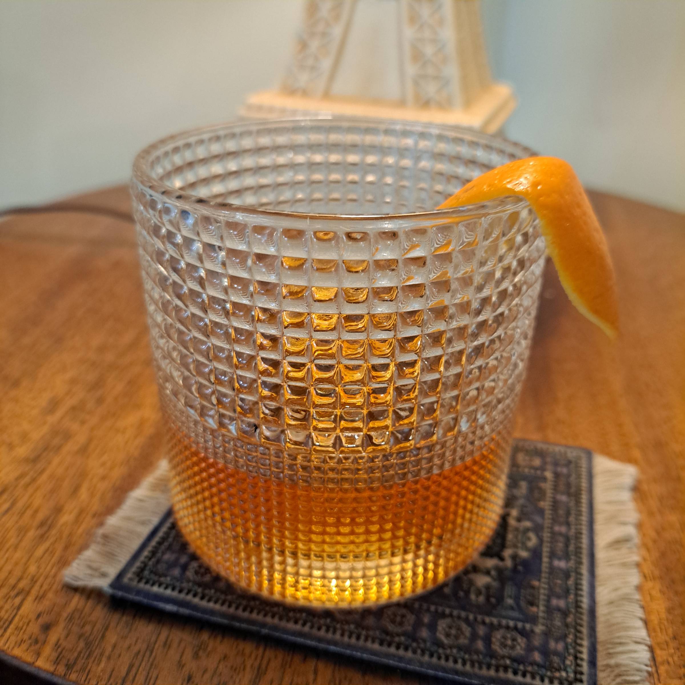 A cocktail glass with an orange peel garnish rests on a coaster on a wood table in front of a lamp