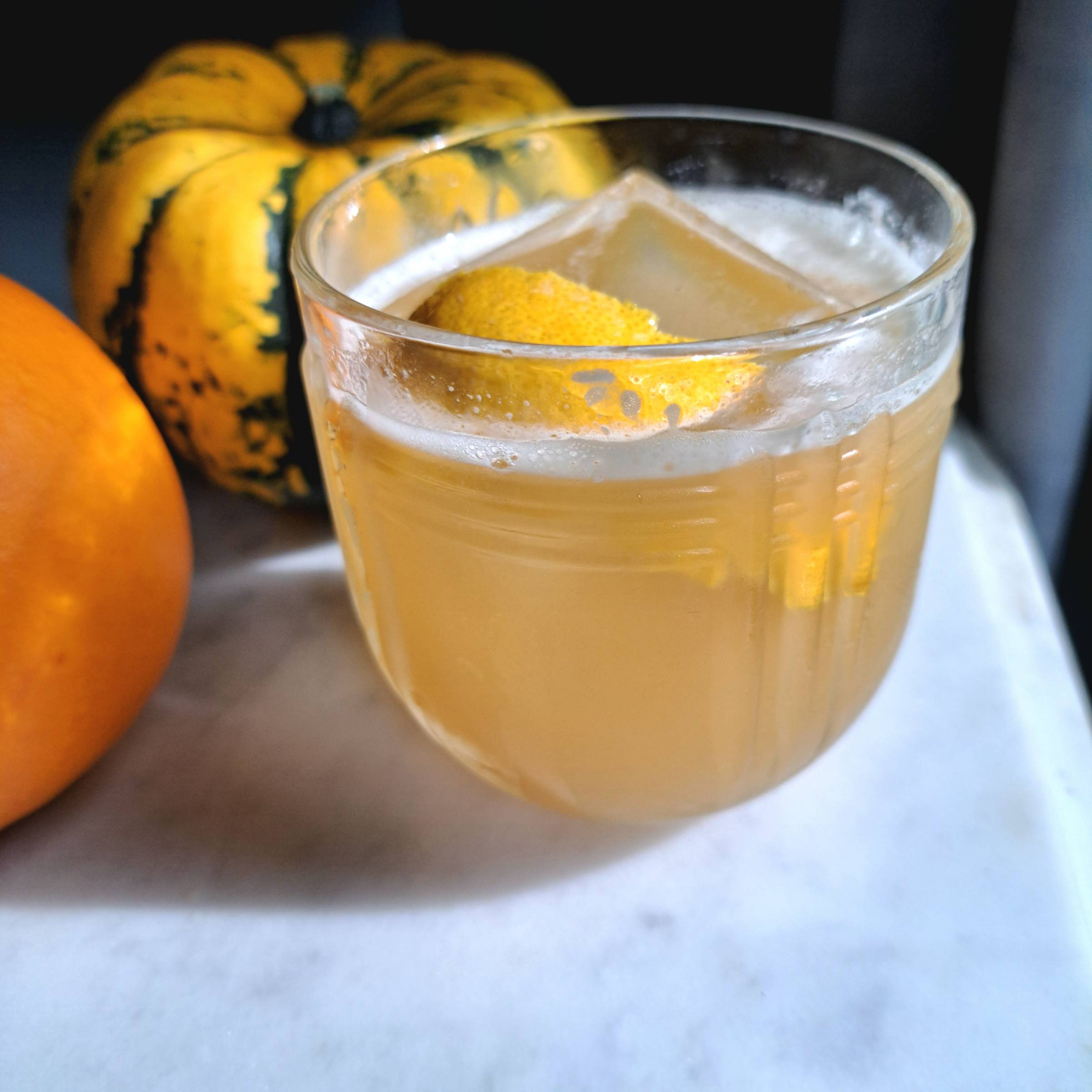 Cardamom Honey Gold Rush bourbon cocktail garnished with a lemon twist and photographed alongside miniature pumpkins for a fall setting.