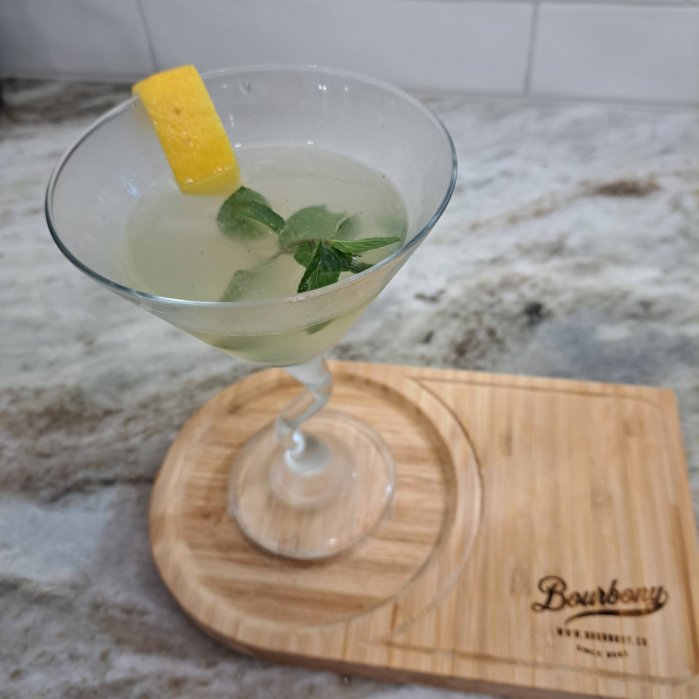 A martini-style glass containing a clear alcoholic beverage with a sprig of mint and a lemon twist sitting on a wooden cutting board