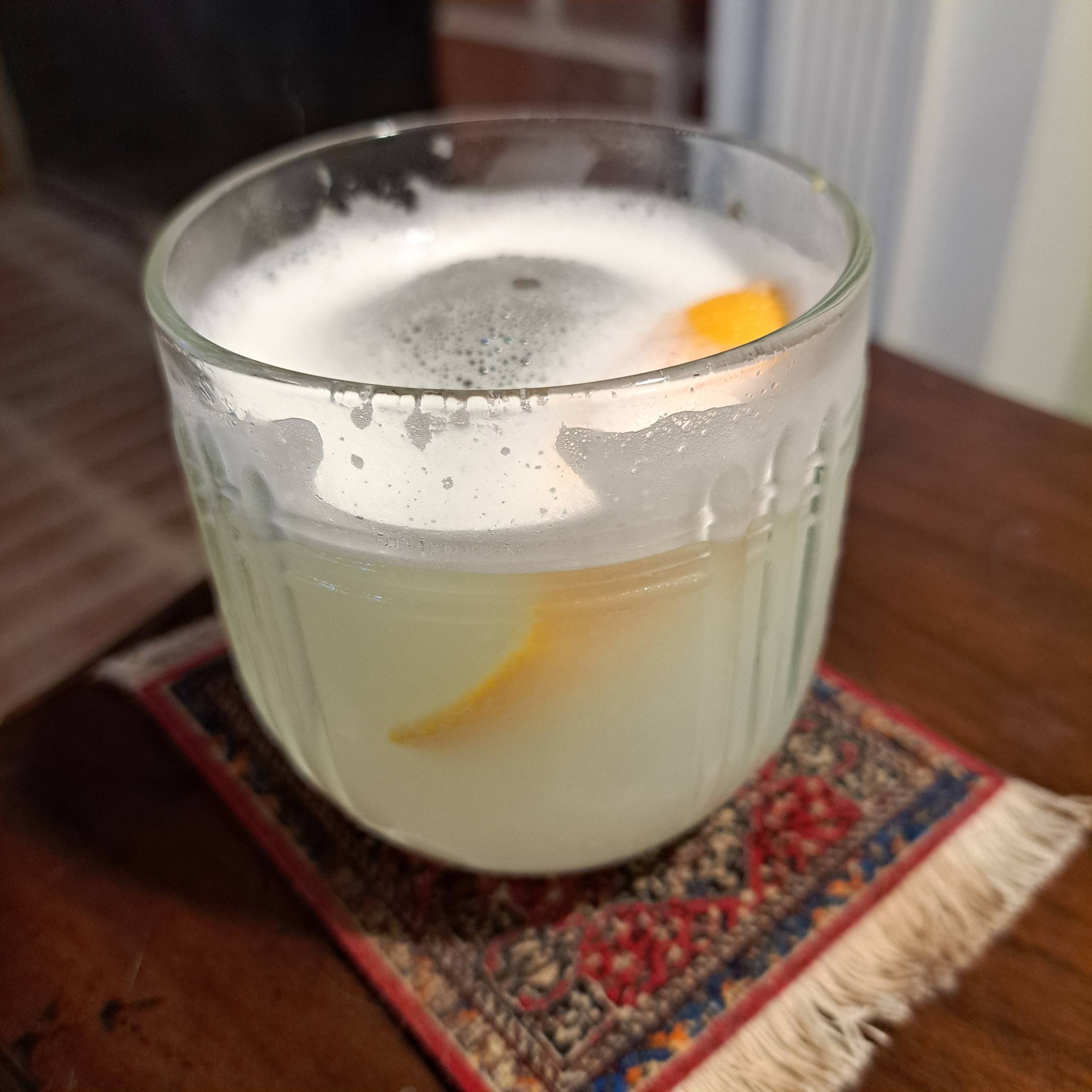 A foamy cocktail with a lemon twist sits on a miniature rug coaster on a wooden tabletop