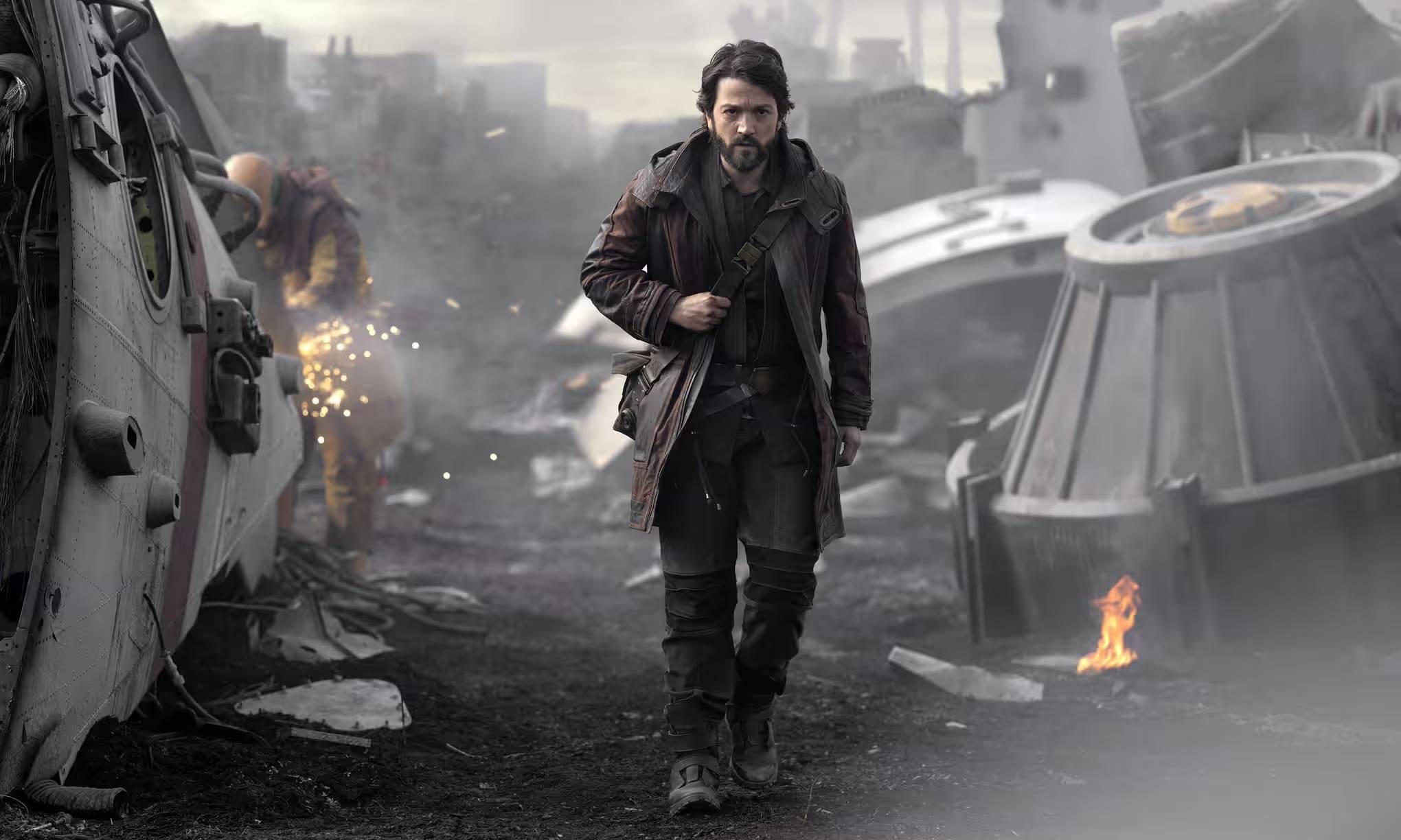 handsome young man in leather jacket walks through flaming wreckage with a determined expression