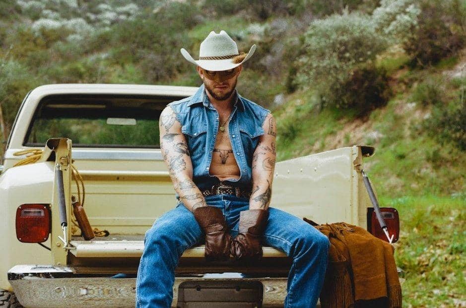sexy cowboy, in sleveless vest sits in the back of a pickup with his legs spread alluringly and posture inviting impure thoughts