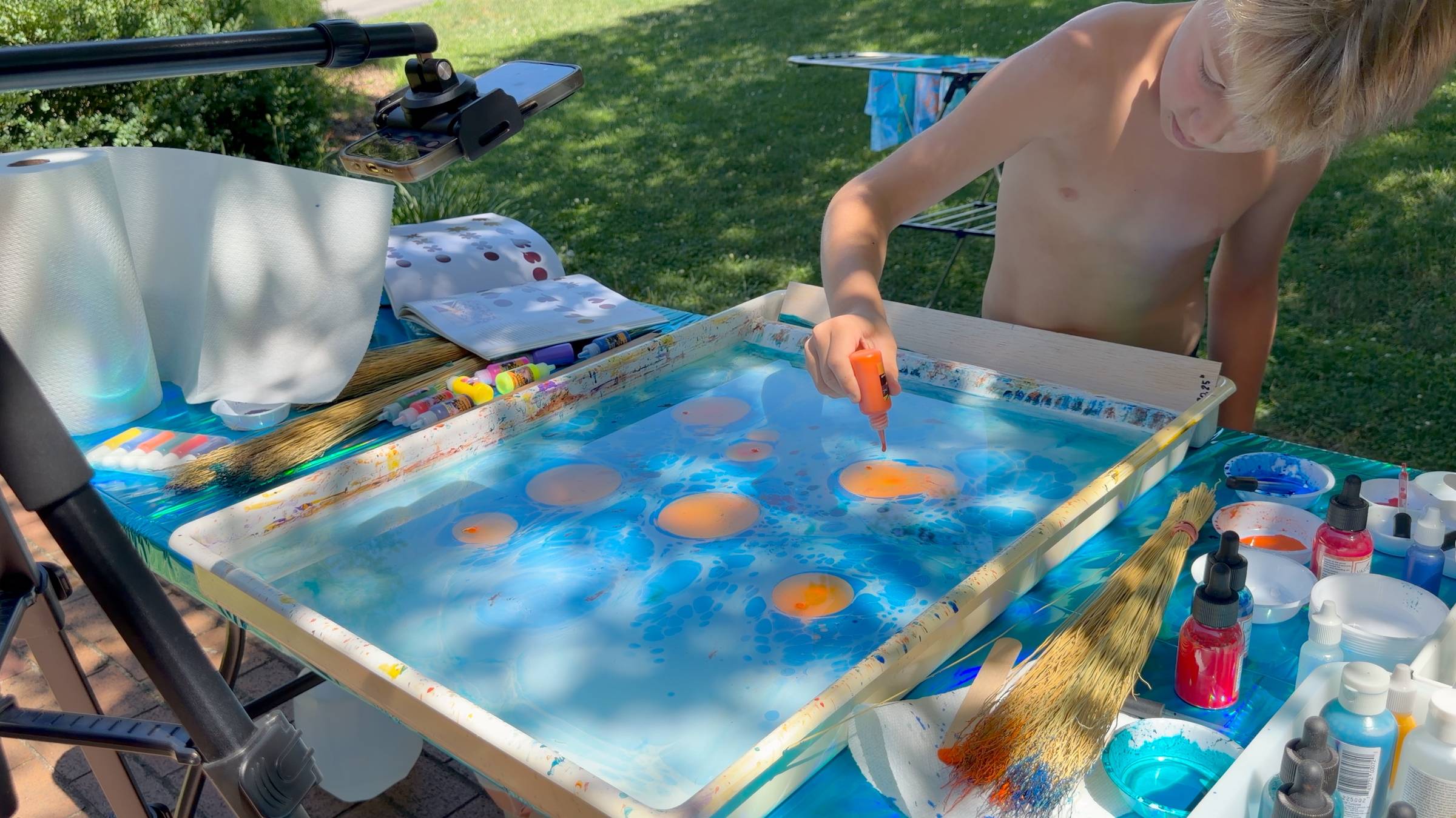 Kody dropping orange paint onto a blue carrageenan size bath — the paint floats on the surface and expands outward in perfect circles. That's surface tension and fluid dynamics happening in real time. Art and physics in one afternoon.