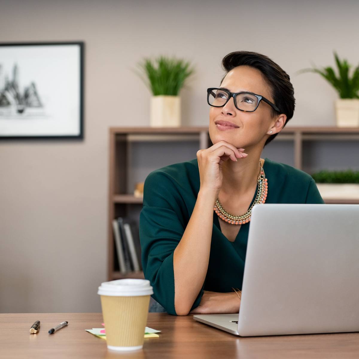 Young Asian Woman in dark green top sitting at laptop head turned, looking up and away, small smile.
