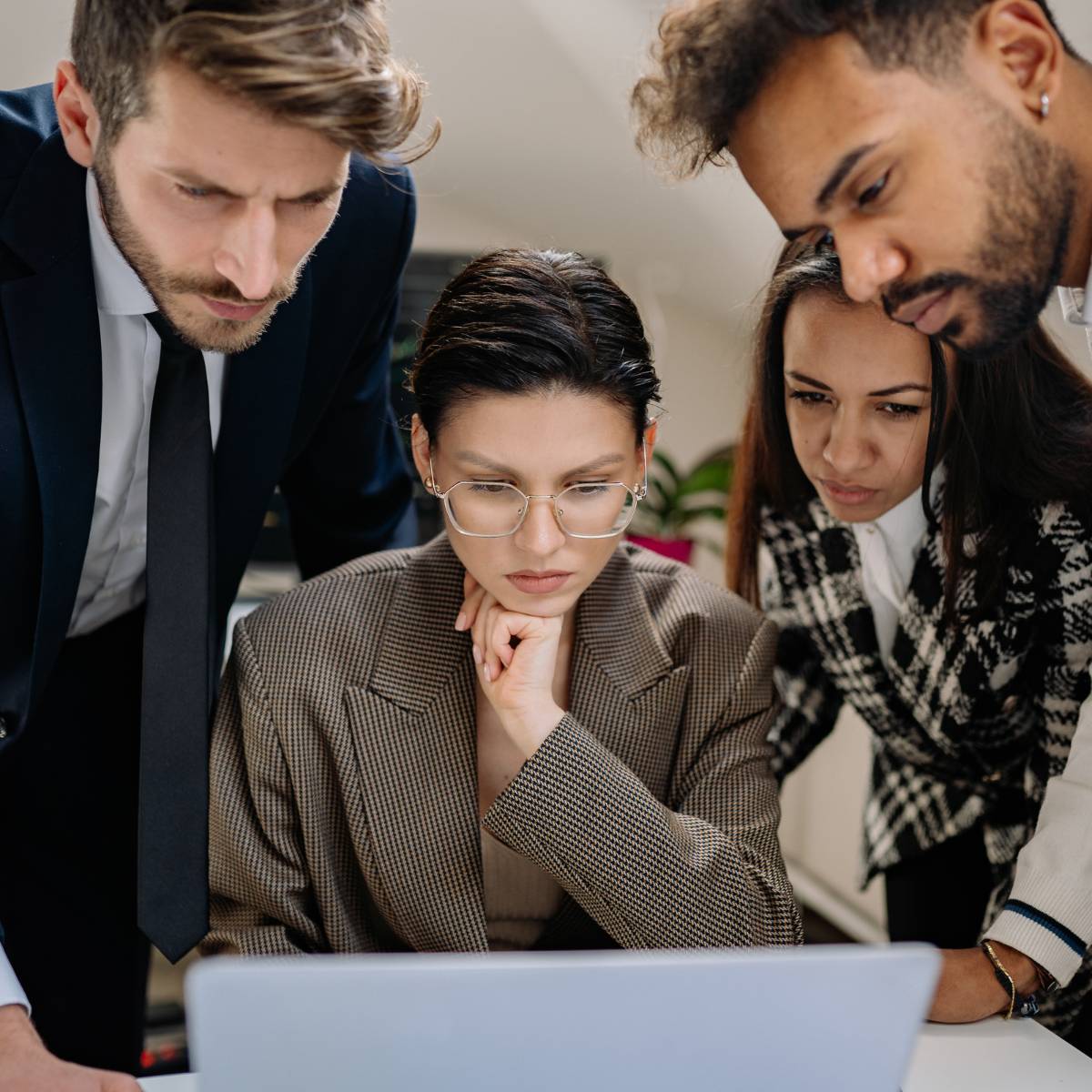 Mature diverse professionals having candid thoughtful discussion, dark-haired woman with gold wire-framed glasses staring into laptop screen, others seriously staring at laptop around her at a table.