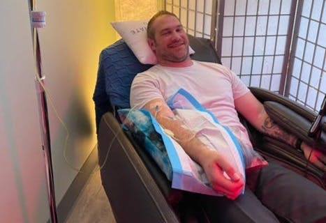 A smiling man with a sleeve tattoo is getting a Crohn's disease infusion of IV nutrients.