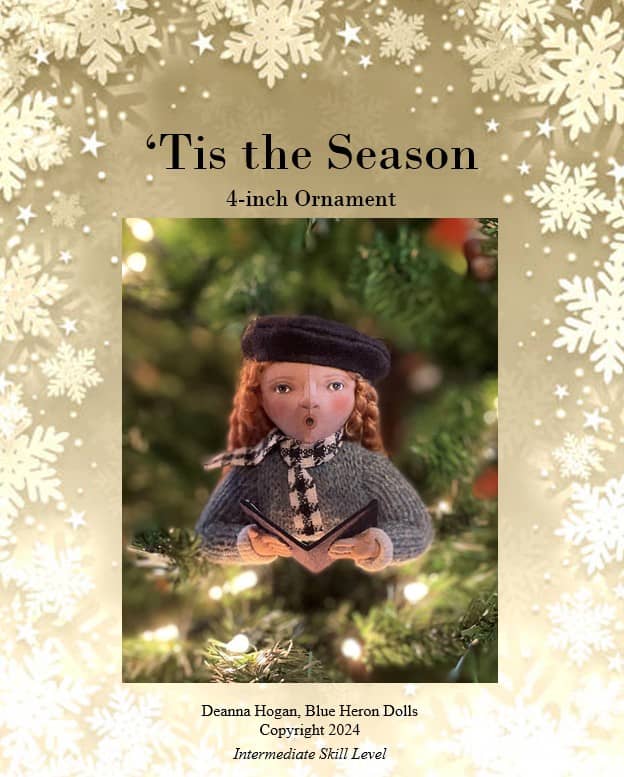 A promotional image for a 4-inch ornament titled "'Tis the Season." The ornament depicts a cute figure wearing a black beret and holding an open book. It has curly, red hair, and is dressed in a knitted sweater and scarf. The background is a blurred Christmas tree with twinkling lights. The border of the image features a pattern of white snowflakes. Text at the bottom credits Deanna Hogan from Blue Heron Dolls and notes "Copyright 2024" with an indication of "Intermediate Skill Level."