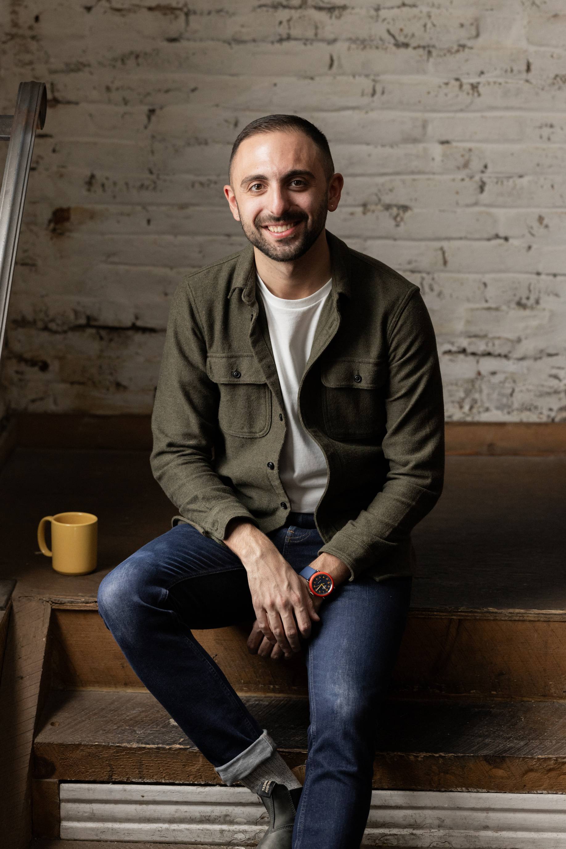 Mike De Socio has short dark hair and a closely-trimmed beard. He is sitting on a wooden staircase in front of a brick wall painted white. A yellow mug sits on the step next to him.