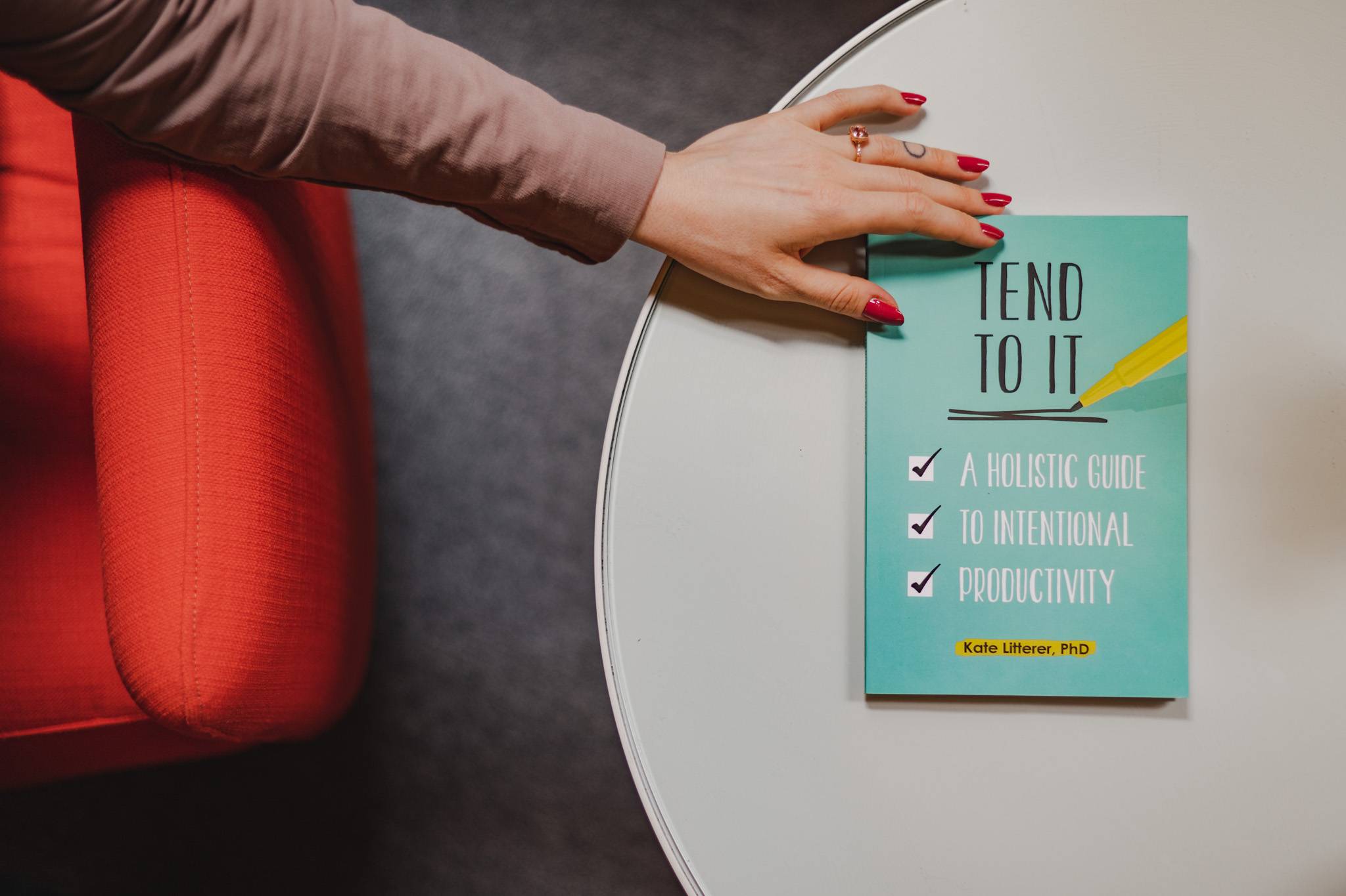 A picture of Tend to It sitting on a table. A hand with bright red nails is reaching toward the book.